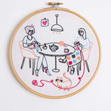 An embroidered picture within an embroidery hoop depicting two women knitting either side of a table with a teapot and cups of tea while a cat plays with the ball of wool on the floor