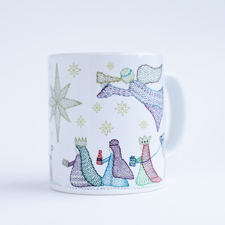 a ceramic mug on a plain back ground. The mug deisgn is of three wise men carrying gifts beneath and angel approaching the Christmas star.  All the features are stitched in a kantha embroidery style in bold colours