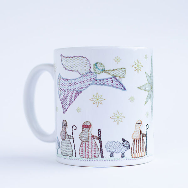 a ceramic mug on a plain back ground. The mug deisgn is of three shepherds and two sheep beneath and angel approaching the Christmas star.  All the features are stitched in a kantha embroidery style in bold colours