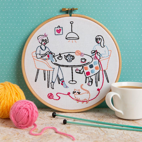 An embroidered picture within an embroidery hoop depicting two women knitting either side of a table with a teapot and cups of tea while a cat plays with the ball of wool on the floor.  The photo include knitting needles, balls of wool and a cup of tea to either side of the embroidery hoop as decoration
