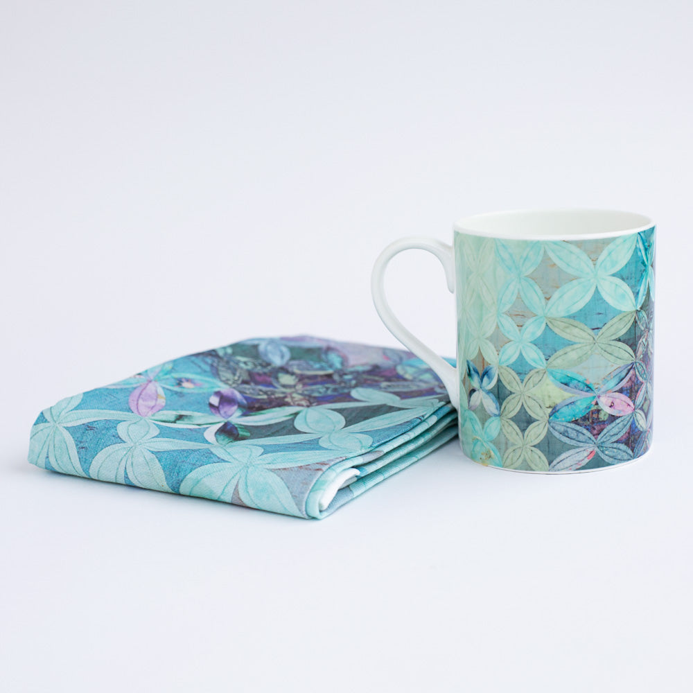 A mug and folded tea towel in a patchwork quilt design in teal , turquoise and purple colour tones